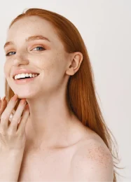 close-up-smiling-redhead-woman-with-pale-skin-freckles-touching-soft-perfect-face-using-skincare-cream-standing-white-wall1